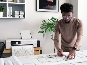Serious Young Black Man In Brown Turtleneck And Glasses Standing At Table And Using Ruler And Pencil While Preparing Technical Drawing